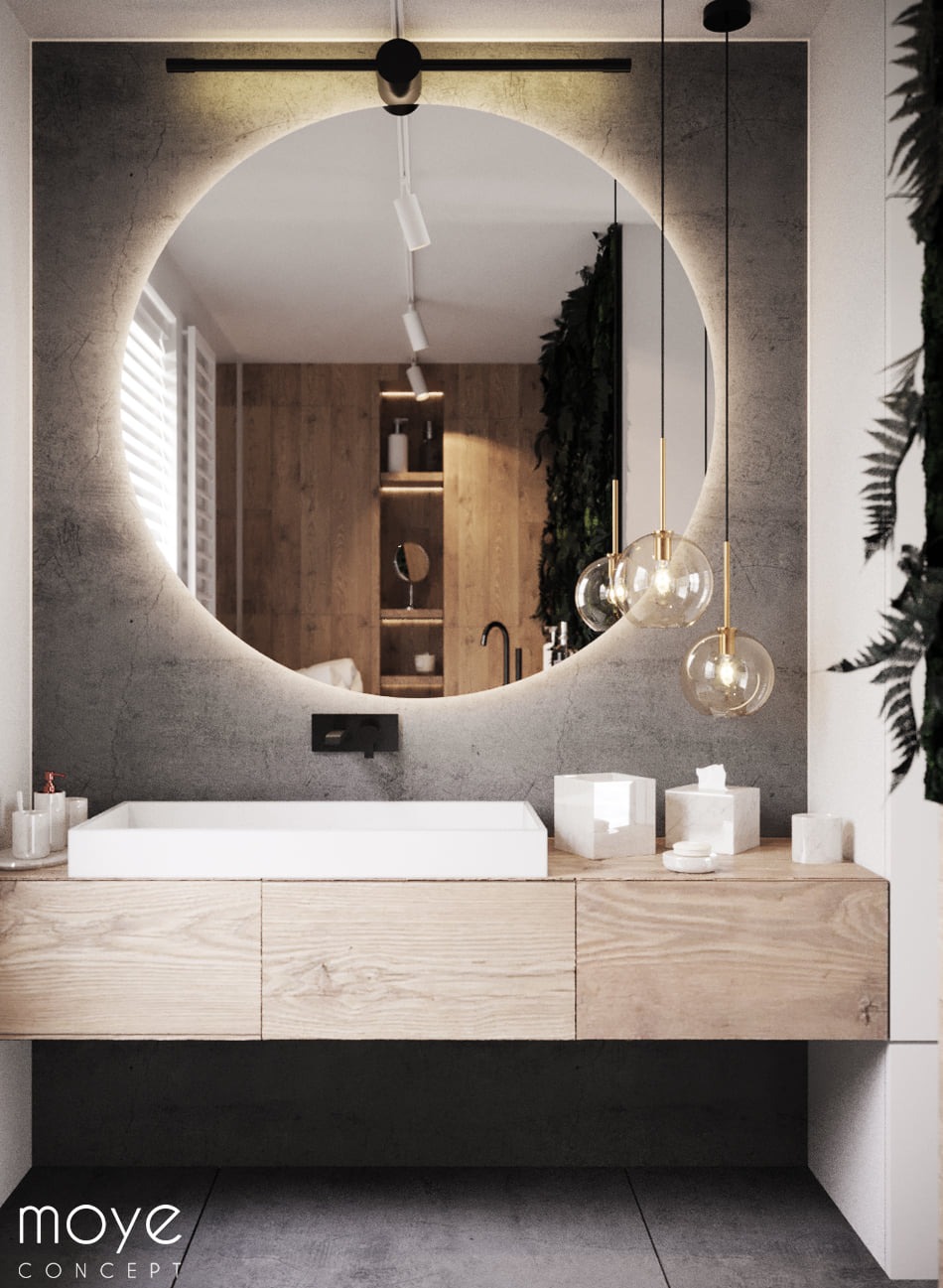 Nowoczesna łazienka z okrągłym lustrem podświetlanym LED, drewnianą szafką pod umywalką, wiszącymi lampami kulami i zieloną ścianą w odbiciu lustra.