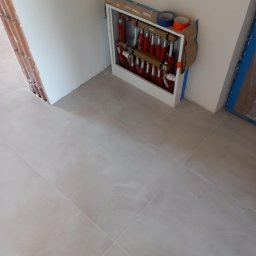 Patryk - Rozdzielacz ogrzewania podłogowego w trakcie montażu, widoczne rury w kolorze czerwonym i białym, taśma malarska i karton zabezpieczający, szare płytki podłogowe.