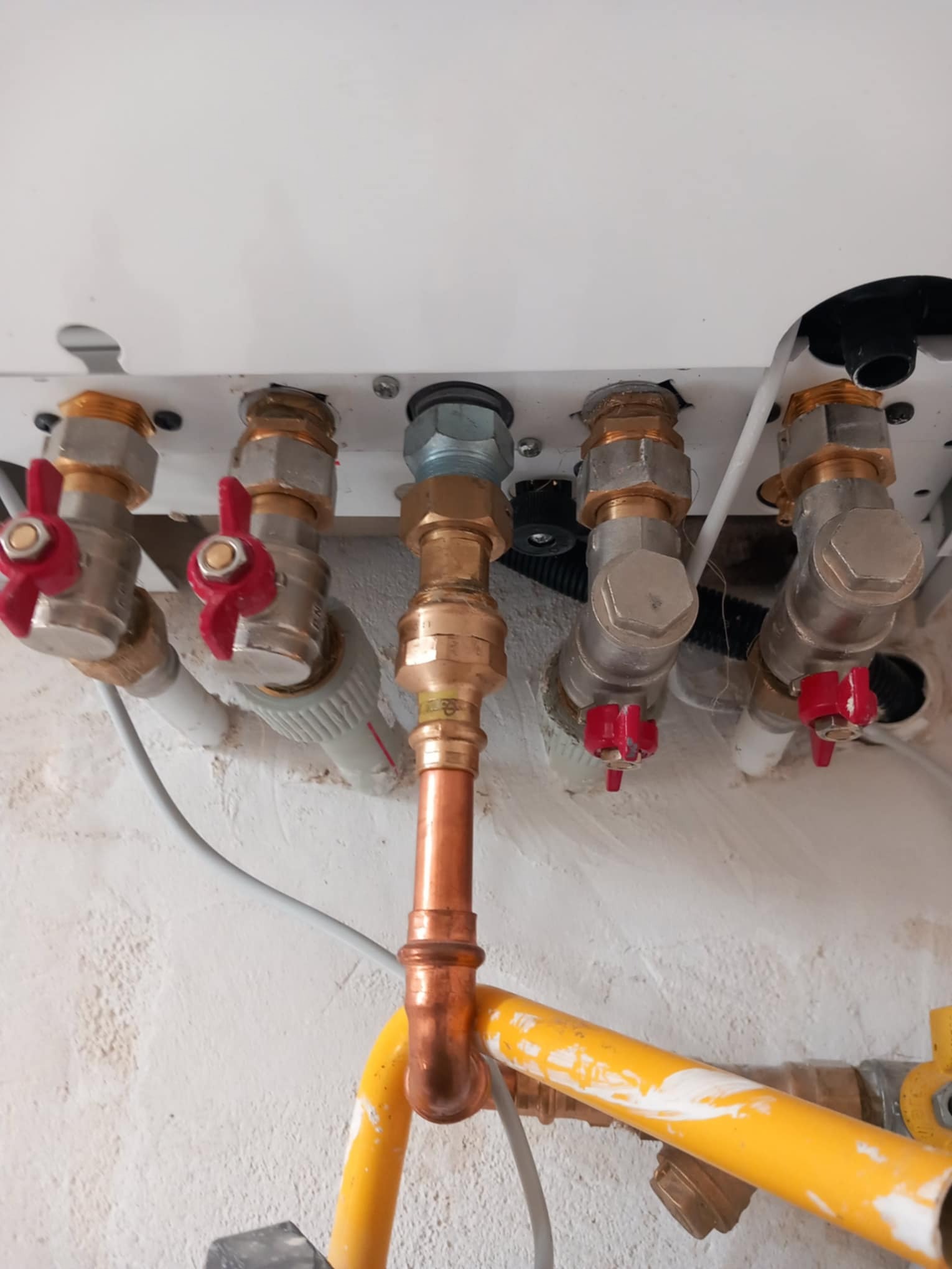 Przyłącze do kotła grzewczego z widocznymi zaworami z czerwonymi pokrętłami, rurami miedzianymi i elementami instalacji gazowej w żółtej osłonie.