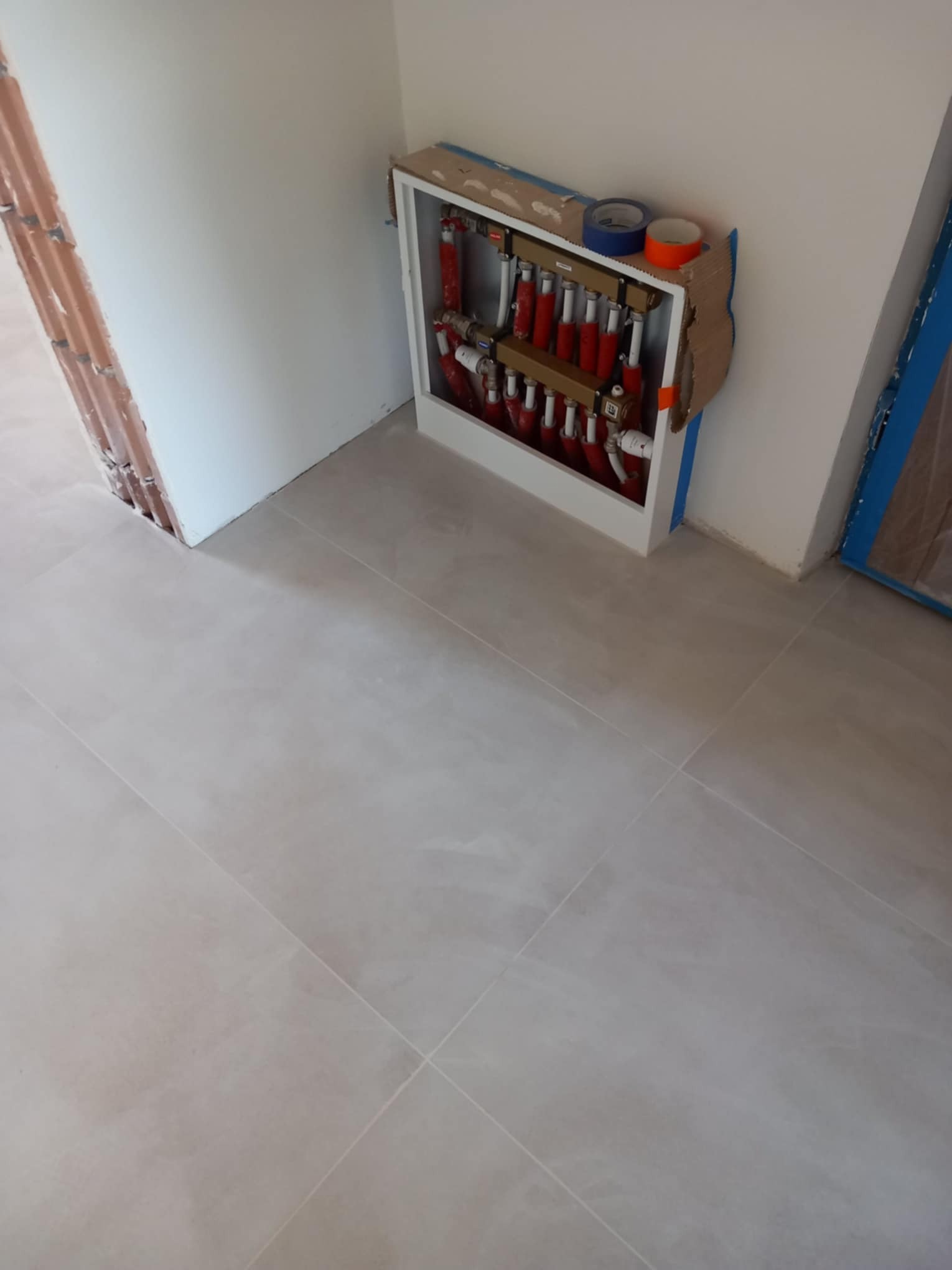 Rozdzielacz ogrzewania podłogowego w trakcie montażu, widoczne rury w kolorze czerwonym i białym, taśma malarska i karton zabezpieczający, szare płytki podłogowe.