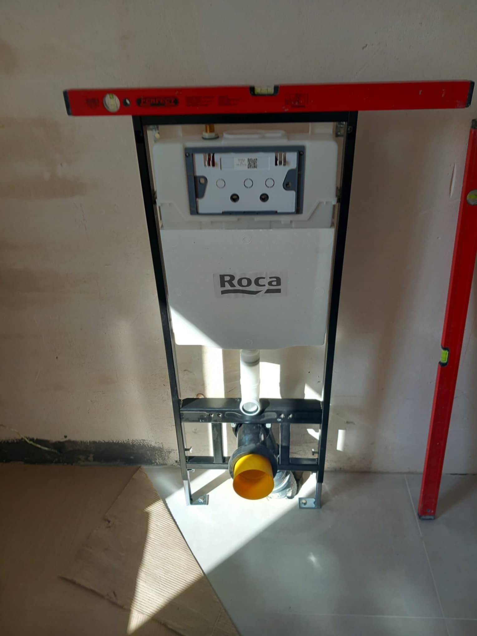 Stelaż podtynkowy WC marki Roca zamontowany w łazience w trakcie prac wykończeniowych, widoczna poziomica dla precyzyjnego montażu i żółta rura odpływowa.