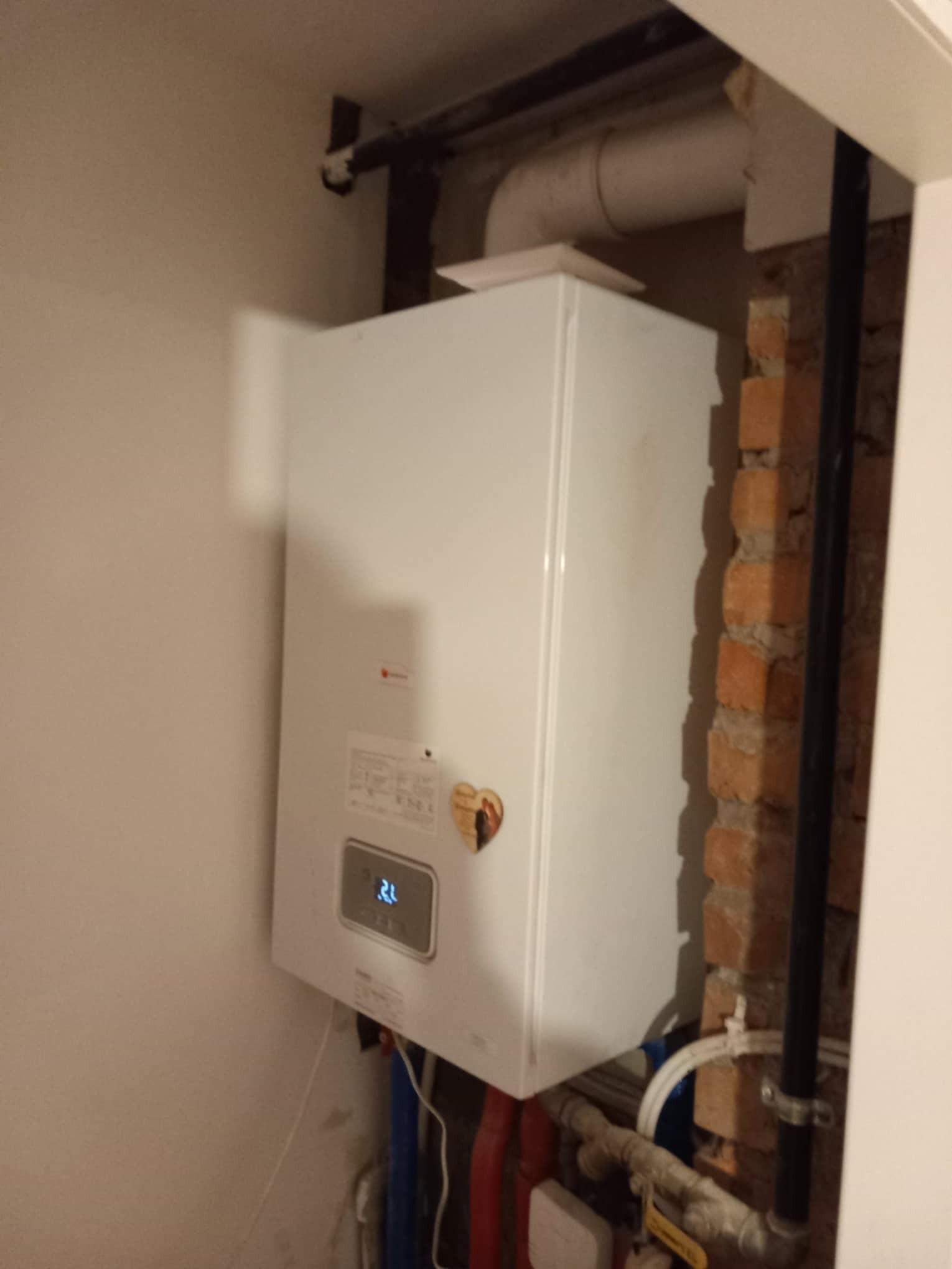 Biały piec gazowy zamontowany na ścianie z widocznymi rurami instalacji, z fragmentem odsłoniętej cegły w tle.