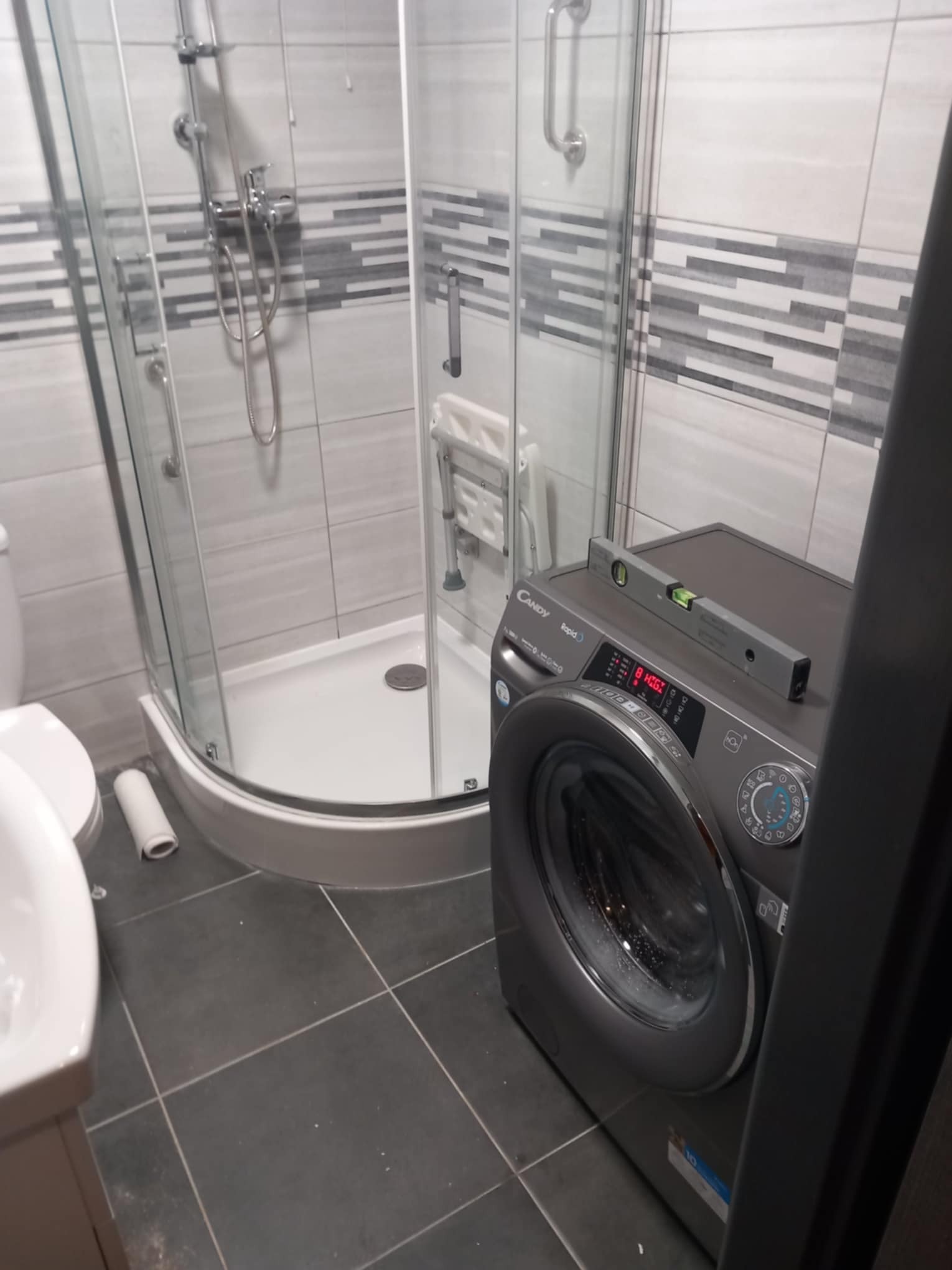 Szara pralka Candy Rapidó ustawiona w rogu łazienki z kabiną prysznicową i umywalką, poziomica na górze pralki, szare płytki podłogowe i ścienne z poziomymi pasami.