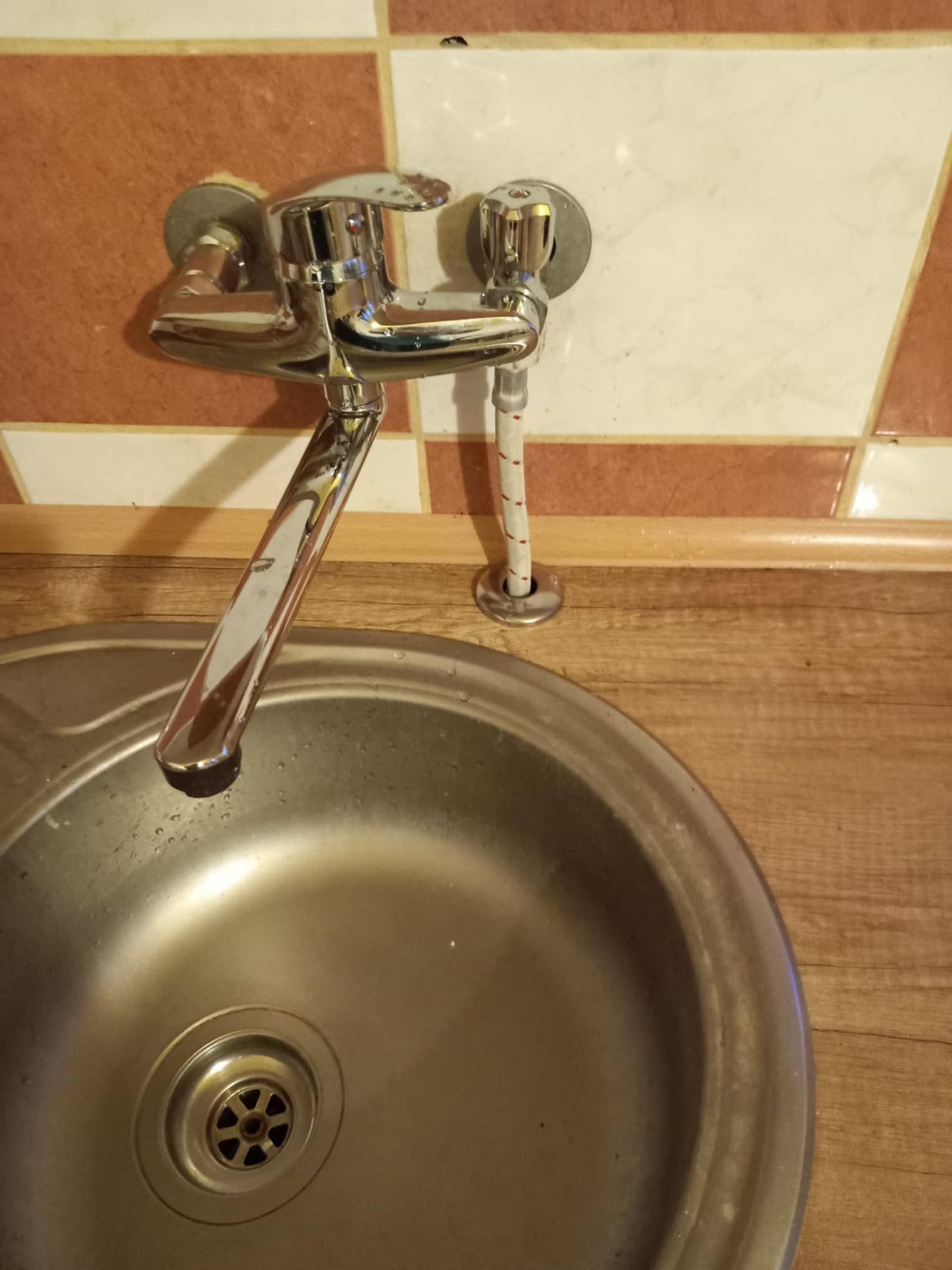 Bateria ścienna chromowana zamontowana nad okrągłym zlewem ze stali nierdzewnej, widoczne przyłącze wężykiem w oplocie.