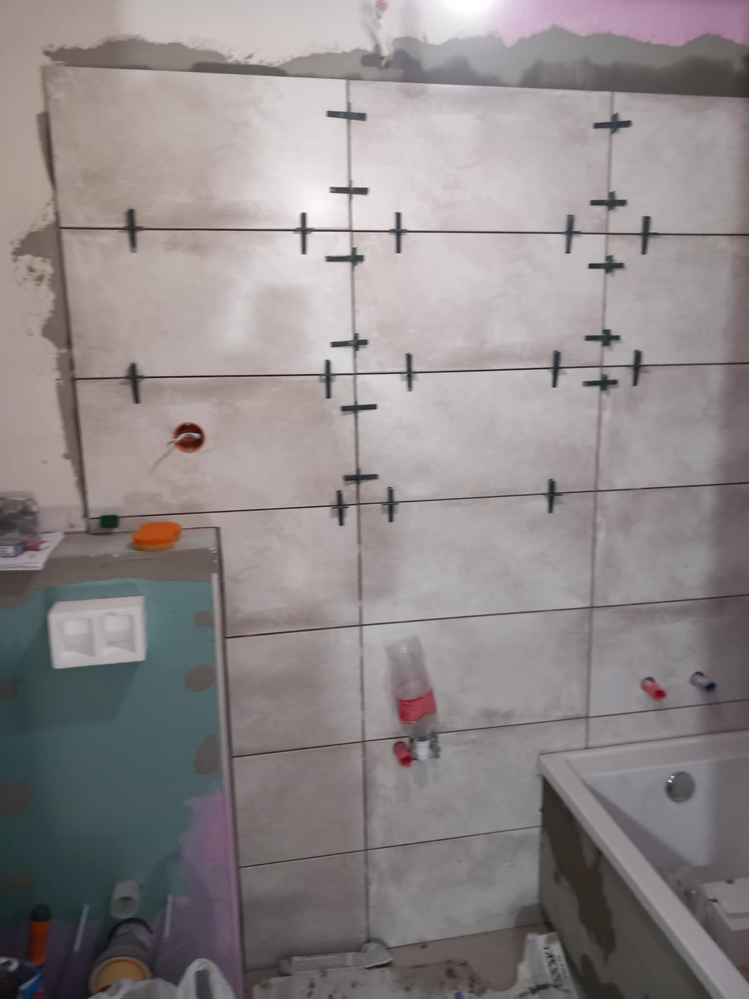 Układanie beżowych płytek na ścianie w łazience, widoczne krzyżyki dystansowe, otwór na gniazdko elektryczne i fragment wanny