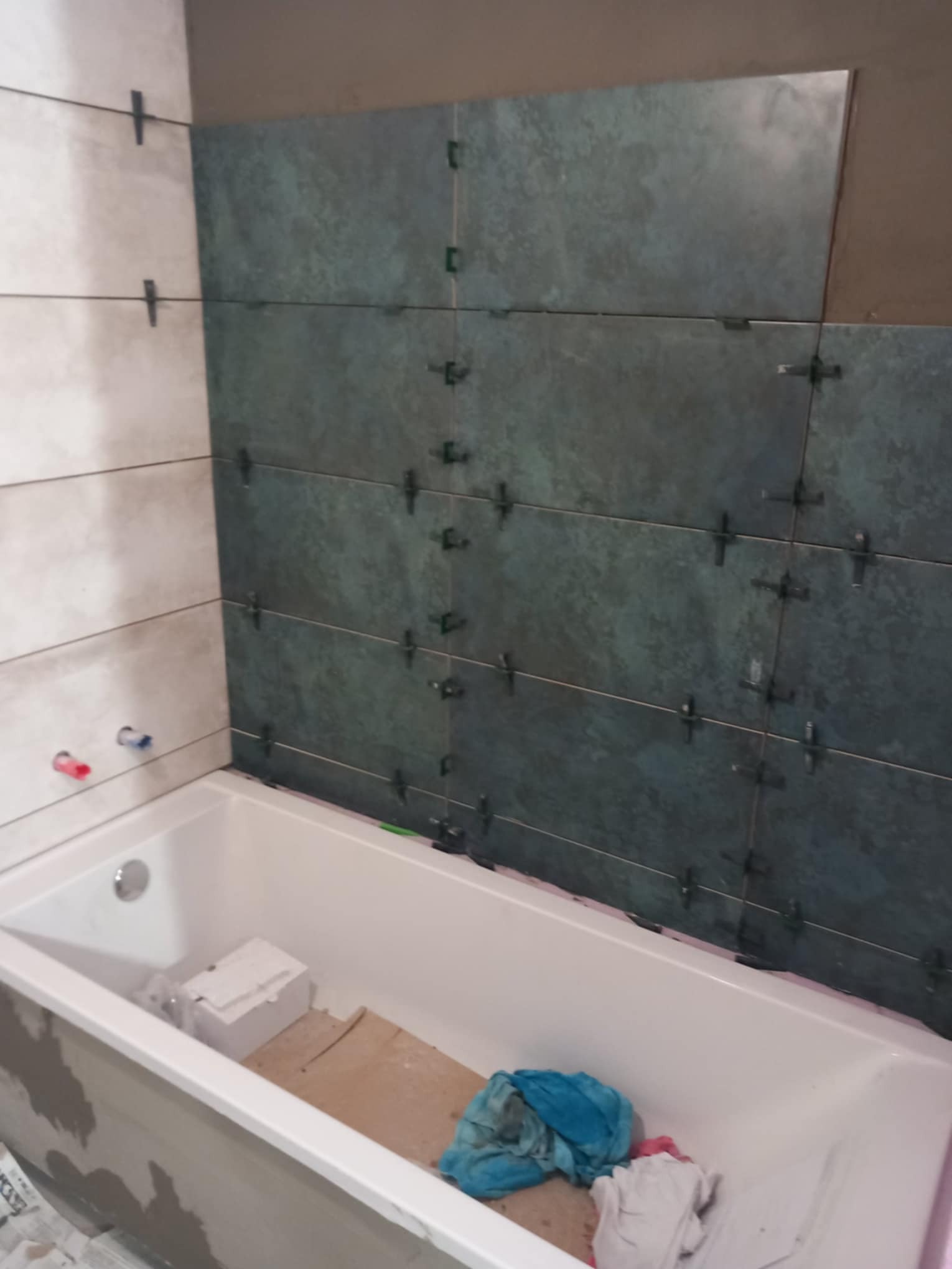 Układanie ciemnozielonych płytek w łazience nad białą wanną, widoczne krzyżyki dystansowe i fragment instalacji wodnej.