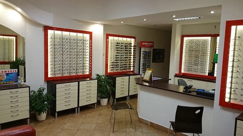 Wnętrze salonu optycznego: ekspozycja okularów w podświetlanych gablotach z czerwonymi ramami, białe komody z szufladami, lada recepcyjna i krzesła dla klientów.