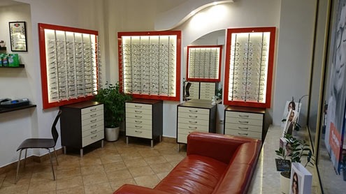 Wnętrze salonu optycznego z ekspozycją okularów w podświetlanych, czerwonych ramkach, komody z szufladami pod ekspozytorami, skórzana kanapa w odcieniach czerwieni i rośliny doniczkowe.