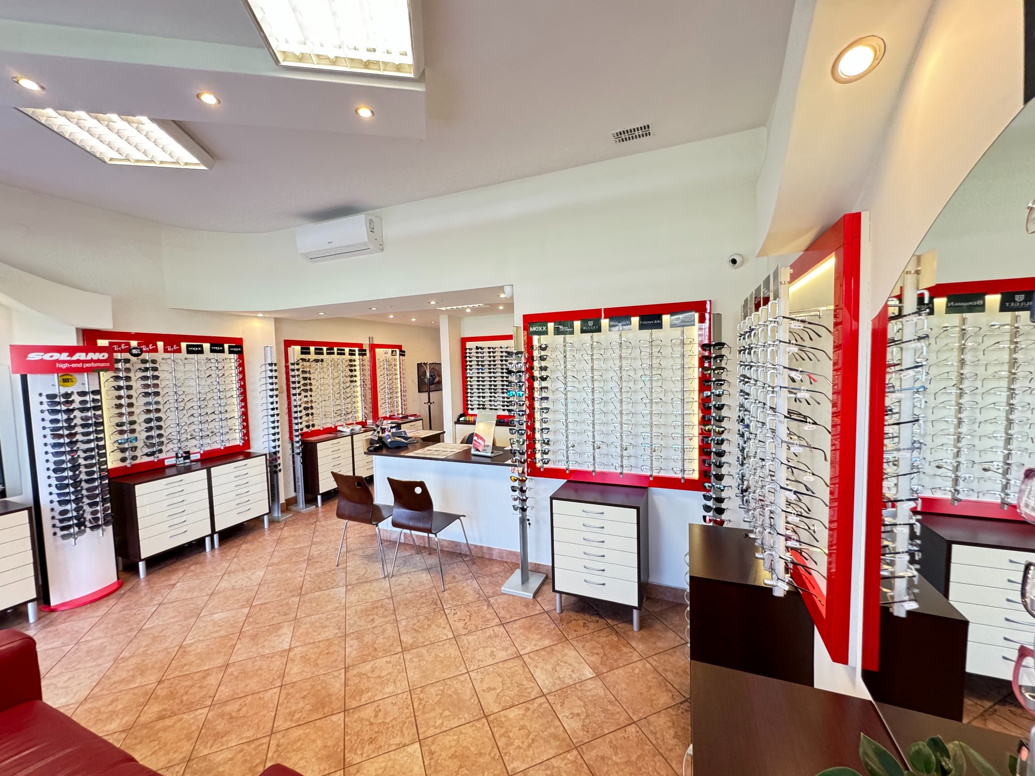 Jasne wnętrze salonu optycznego z ekspozycją okularów na ścianach i szafkach, widoczne krzesła dla klientów i stanowisko obsługi.