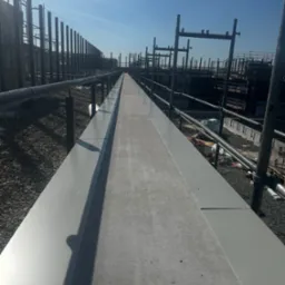 Długi, płaski dach z szarej blachy, widok z góry w perspektywie zbieżnej. W tle konstrukcja szkieletowa budynku i błękitne niebo.