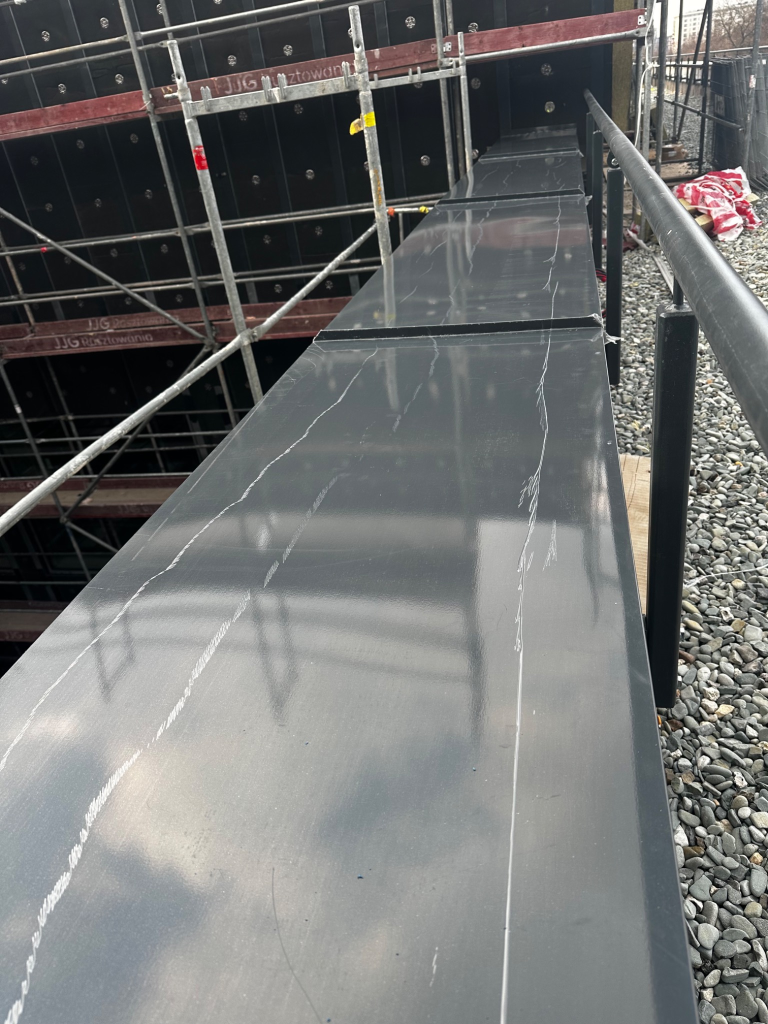 Szare panele blaszane z białymi śladami po zarysowaniach, ułożone na rusztowaniu, widok z góry na tle żwirowego podłoża.