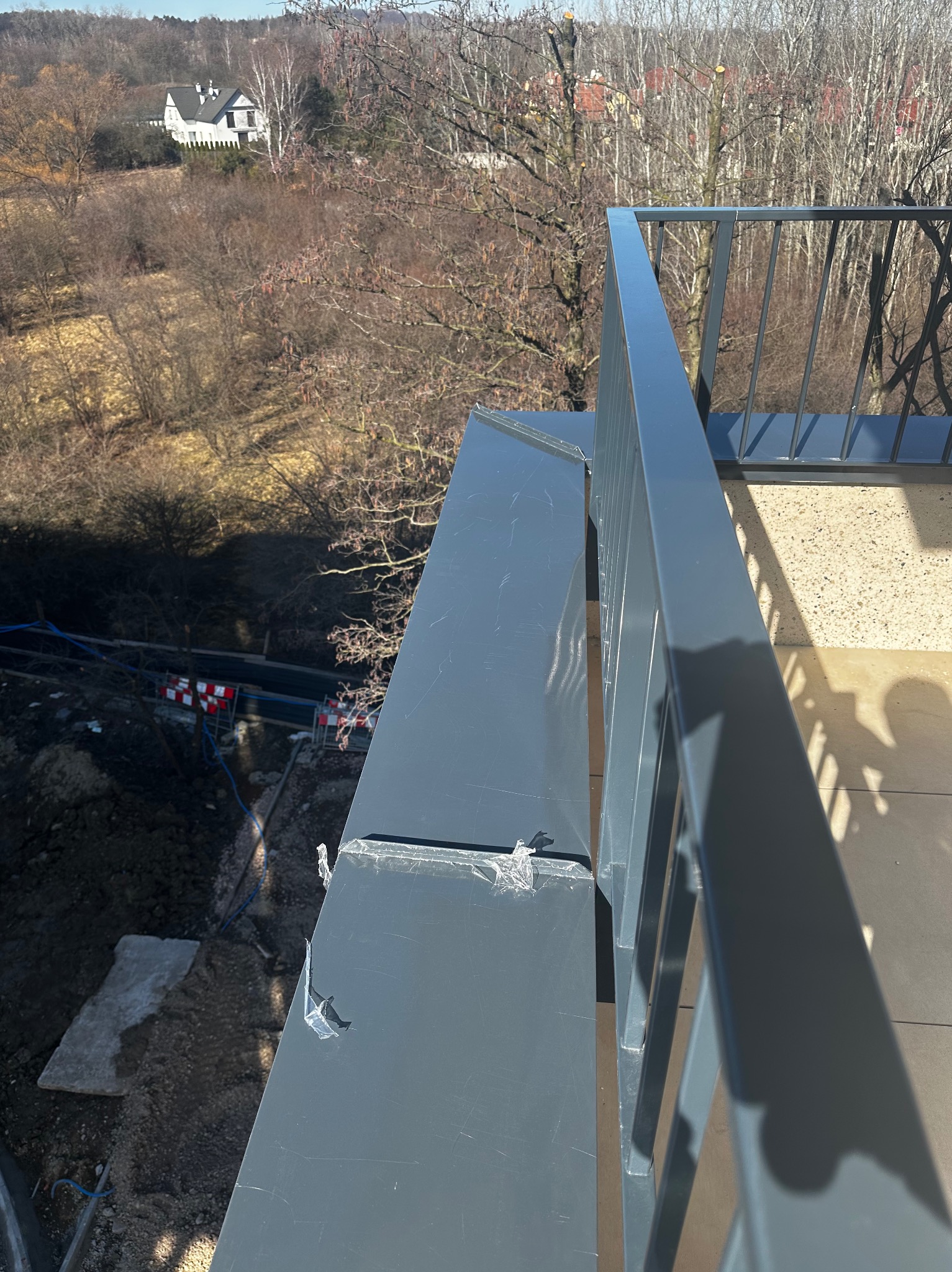 Szara obróbka blacharska na balkonie z widocznymi śladami folii ochronnej, na tle jesiennego krajobrazu i białego domu.