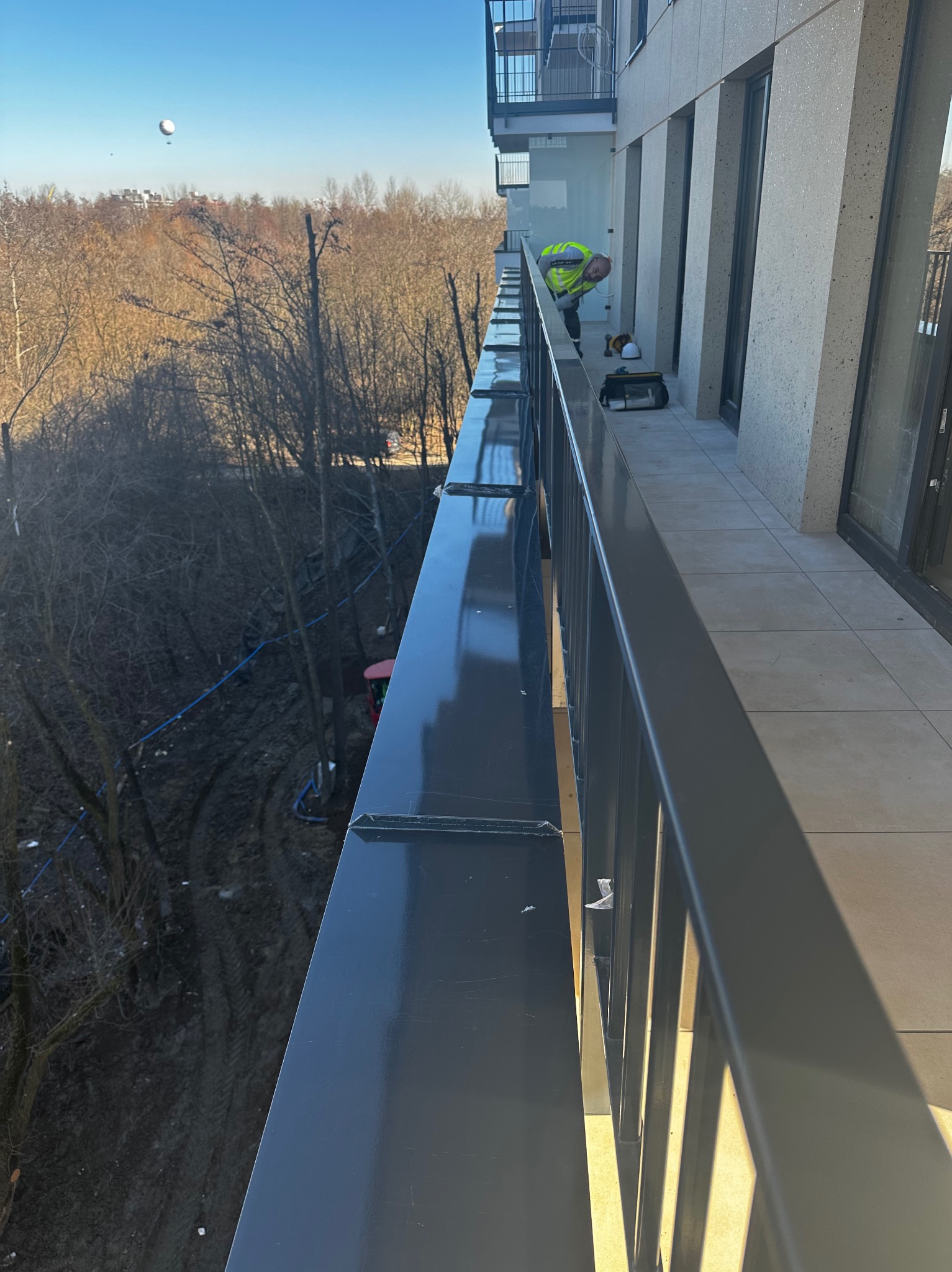 Pracownik w żółtej kamizelce, pochylony nad metalową konstrukcją balkonu w Krakowie, prawdopodobnie przy montażu lub wykończeniu elementów. W tle widoczne drzewa i budynki.