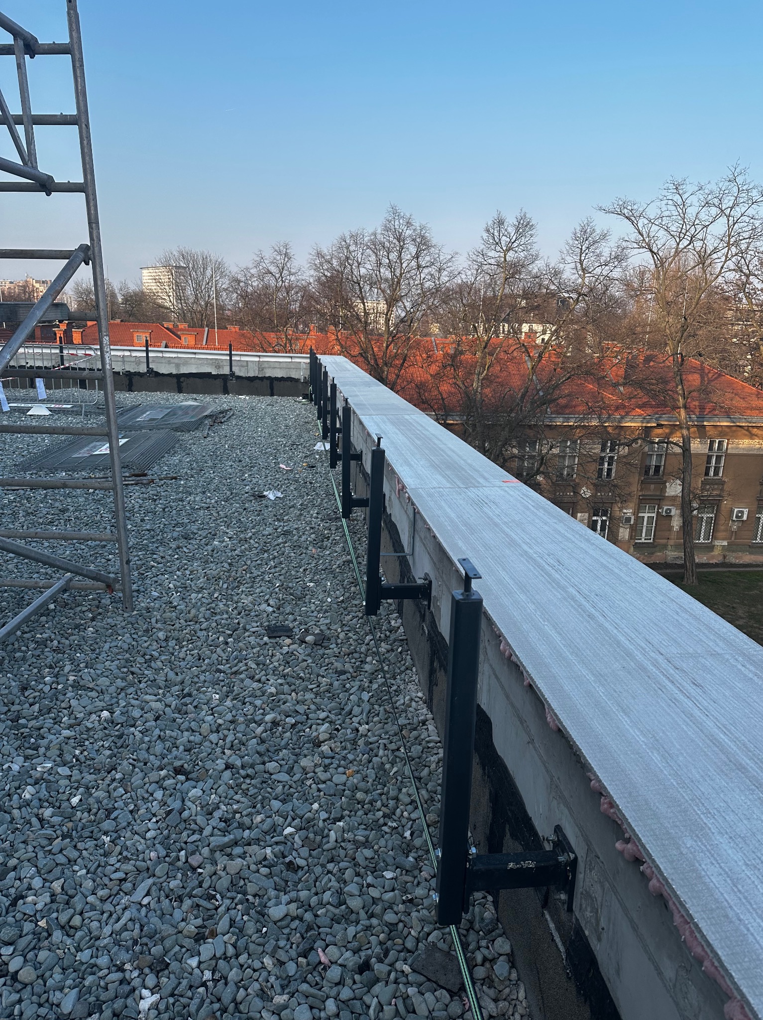 Widok z góry na fragment dachu pokrytego drobnym żwirem, z zamontowanymi czarnymi wspornikami przy krawędzi, w tle budynki z czerwonymi dachami i drzewa bez liści.