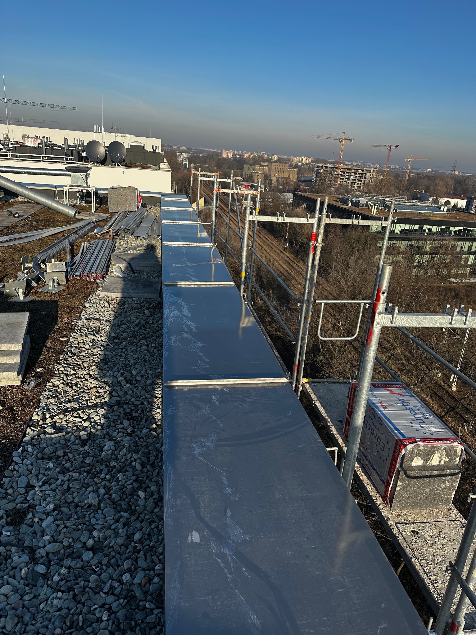 Nowe, błyszczące elementy blaszane na dachu budynku w trakcie montażu, widoczne rusztowania i cień osoby wykonującej prace, w tle panorama miasta z dźwigami budowlanymi.