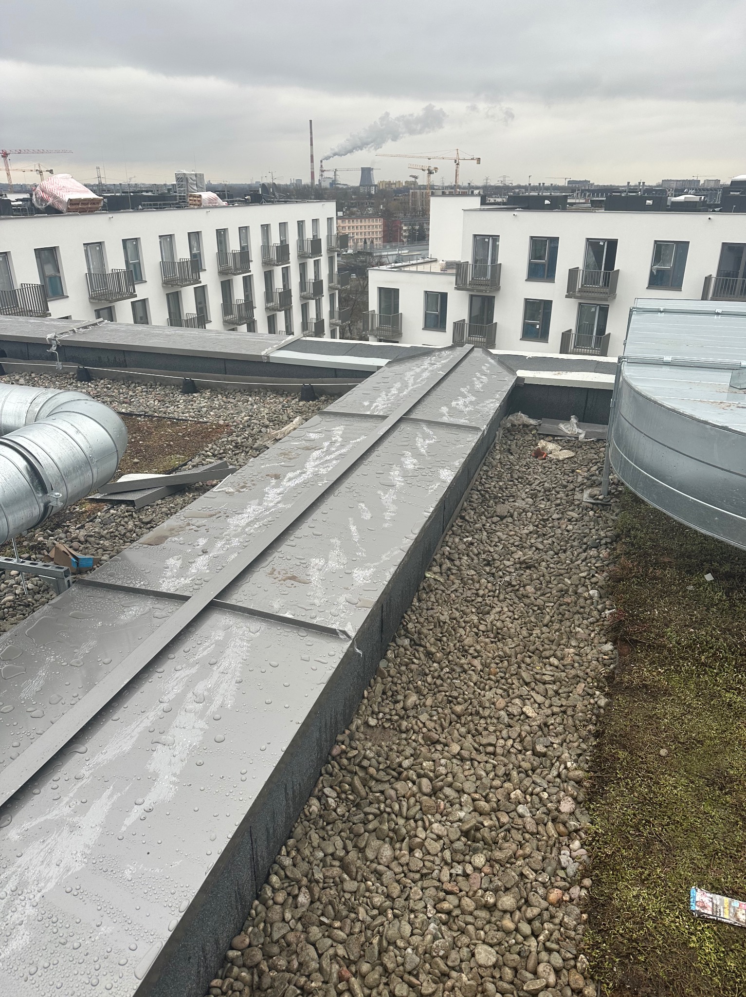 Widok z góry na płaski dach budynku z szarymi panelami na żwirze, widoczne elementy wentylacji i budynki mieszkalne w tle w pochmurny dzień.