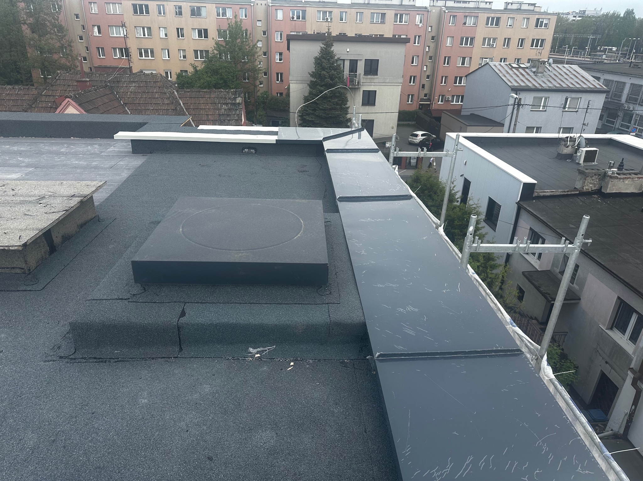 Wykończony, płaski dach pokryty papą termozgrzewalną z obróbkami blacharskimi w kolorze grafitowym. Widok z góry na tle zabudowy miejskiej w Krakowie.