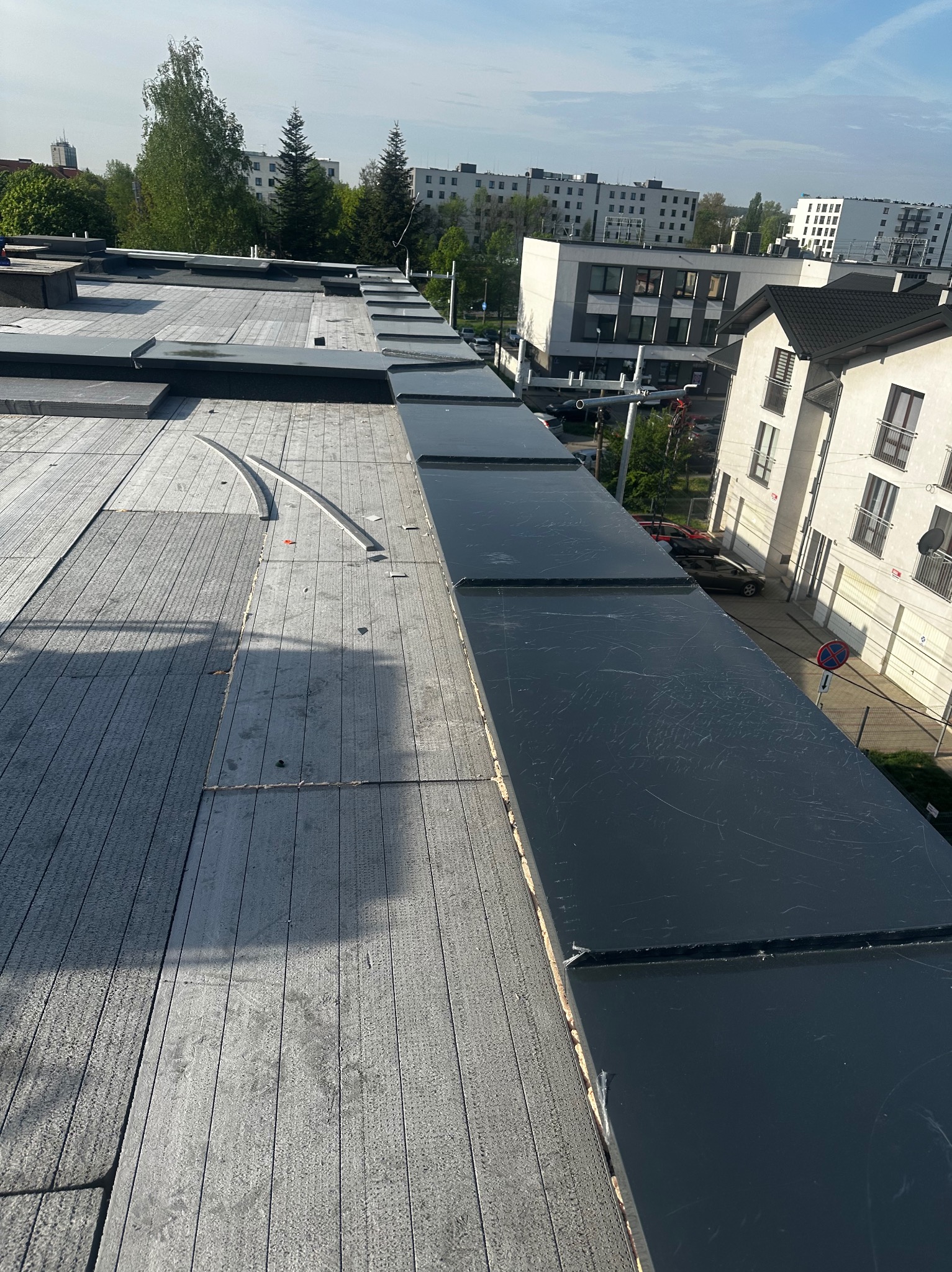 Widok z góry na fragment dachu z szarą, drewnopodobną powierzchnią i czarnymi panelami. W tle budynki mieszkalne i drzewa.