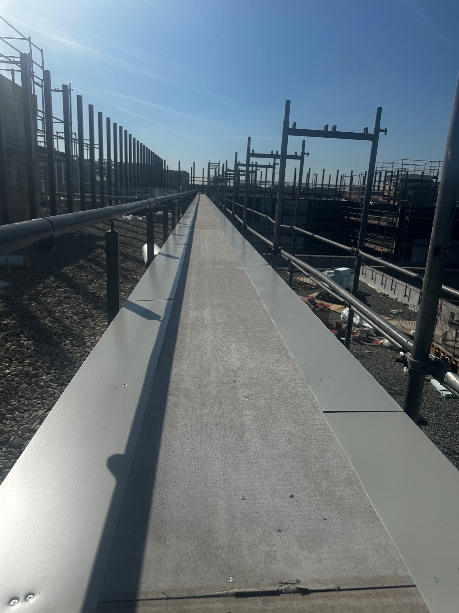 Długi, płaski dach z szarej blachy, widok z góry w perspektywie zbieżnej. W tle konstrukcja szkieletowa budynku i błękitne niebo.