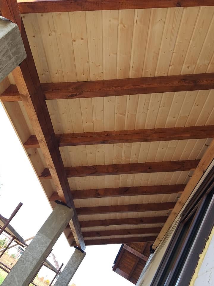 Drewniany podbitka dachowa z belek, widok z dołu, jasne panele z widocznymi słojami drewna, bejcowane na brązowo belki, betonowe filary podtrzymujące konstrukcję.