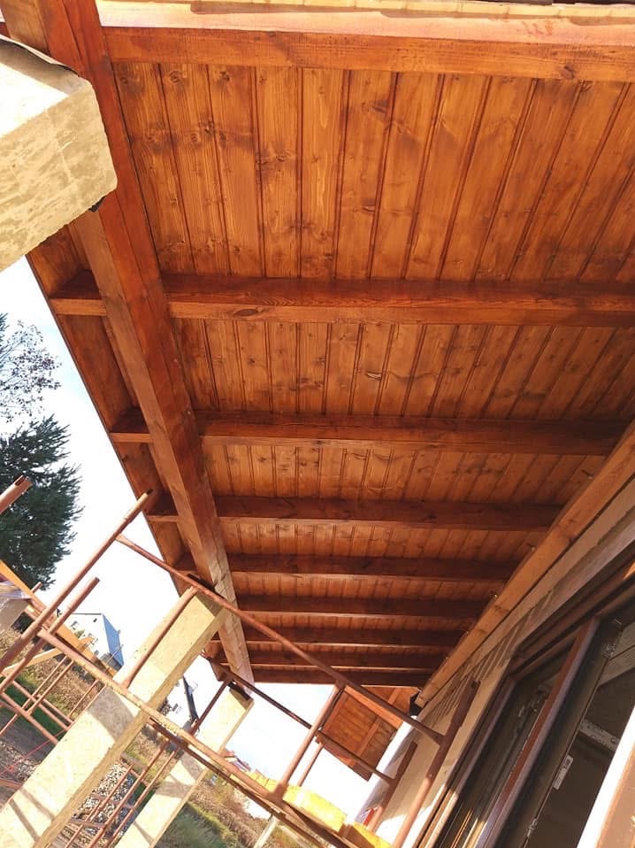 Drewniana podbitka dachowa z belek, świeżo po impregnacji, widoczna spod spodu; elementy konstrukcyjne w trakcie montażu.