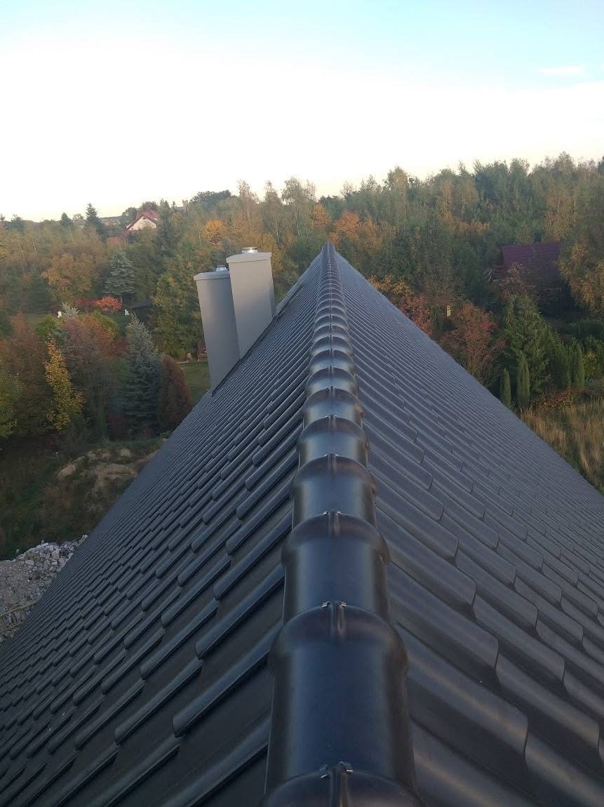 Ciemna, metalowa dachówka ceramiczna z rynną kalenicową, widziana z góry pod kątem. W tle drzewa i kominy wentylacyjne.