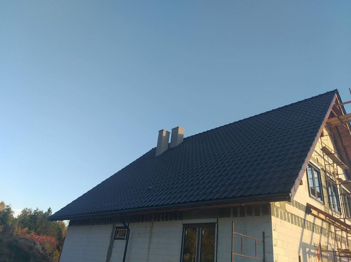 Ciemna dachówka na skośnym dachu domu w budowie, widoczne dwa kominy i rusztowanie. Ujęcie od dołu, błękitne niebo w tle.