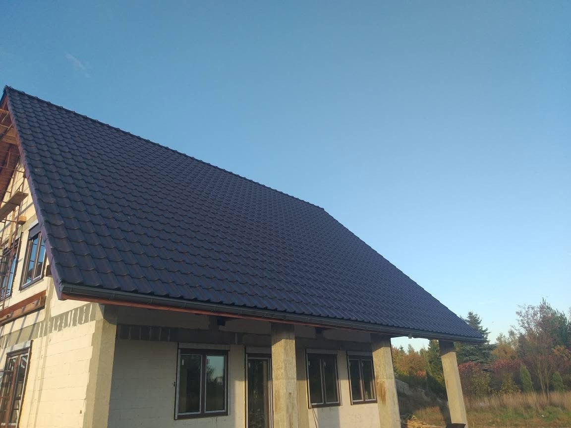 Niebieski dach z dachówki ceramicznej na surowym budynku w trakcie budowy, widok z dołu w słoneczny dzień. Widoczne rynny i fragmenty konstrukcji dachu.