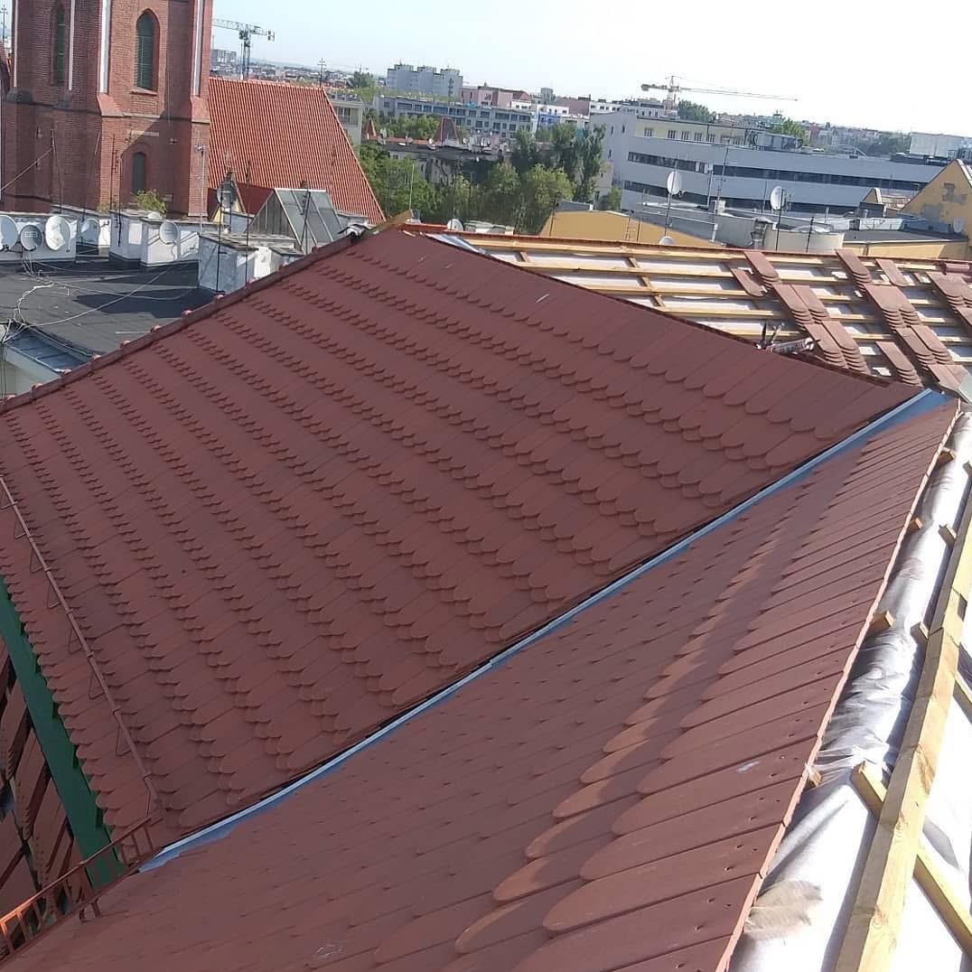 Częściowo odnowiony dach z brązową dachówką karpiówką na tle miejskiej zabudowy z kościołem i żurawiem budowlanym. Widoczny fragment konstrukcji więźby dachowej.
