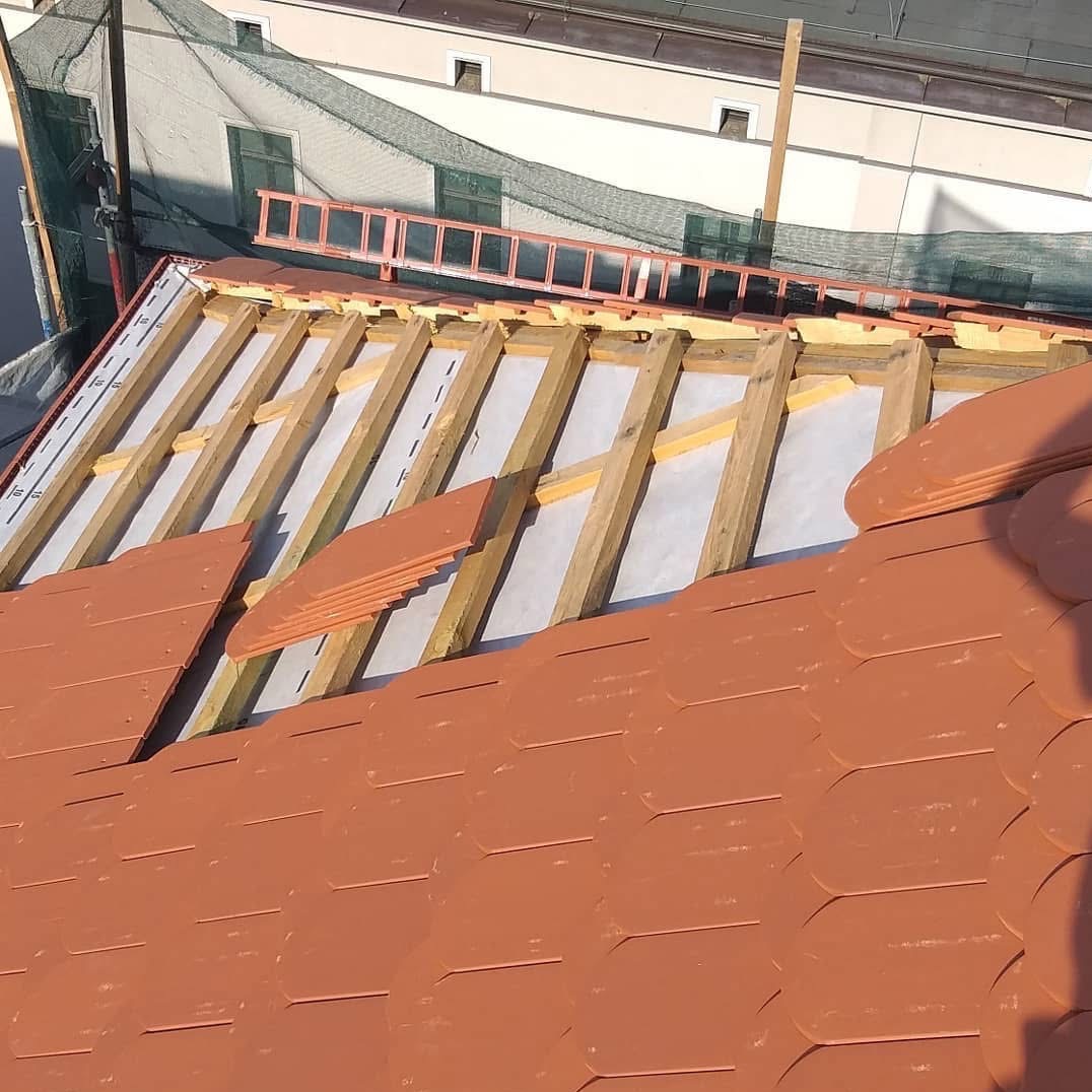 Częściowo pokryty dach z czerwoną dachówką w kształcie łuski, widoczne drewniane elementy konstrukcji i drabina w tle. Prace dekarskie w toku, widok z góry na konstrukcję.