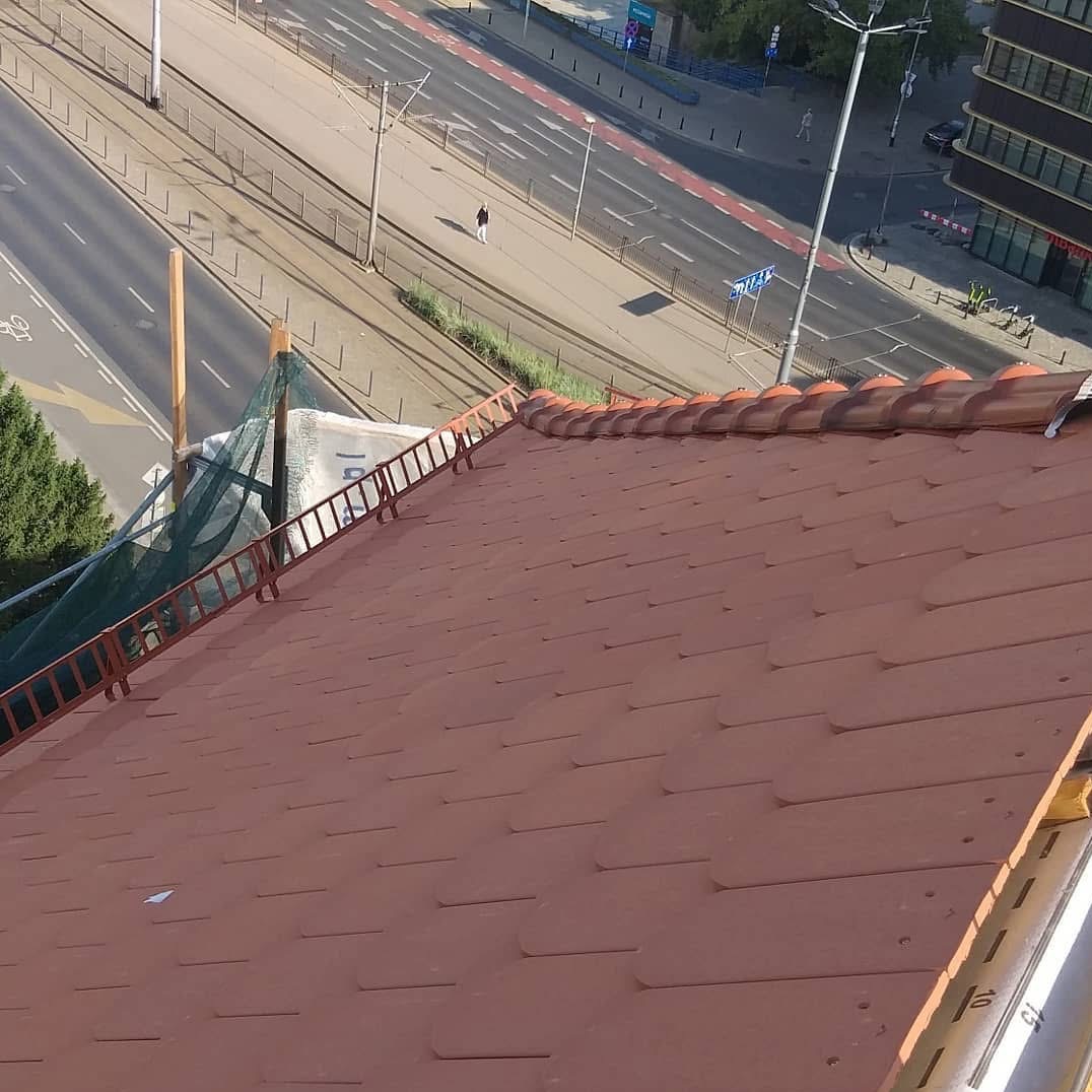 Czerwona dachówka ceramiczna z zamontowaną drabiną dekarską na tle miejskiego krajobrazu z ulicą i budynkami, widok z góry.