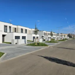 Nowoczesne osiedle domów szeregowych z płaskimi dachami, szarymi bramami garażowymi i kostką brukową na podjazdach, sfotografowane w słoneczny dzień.