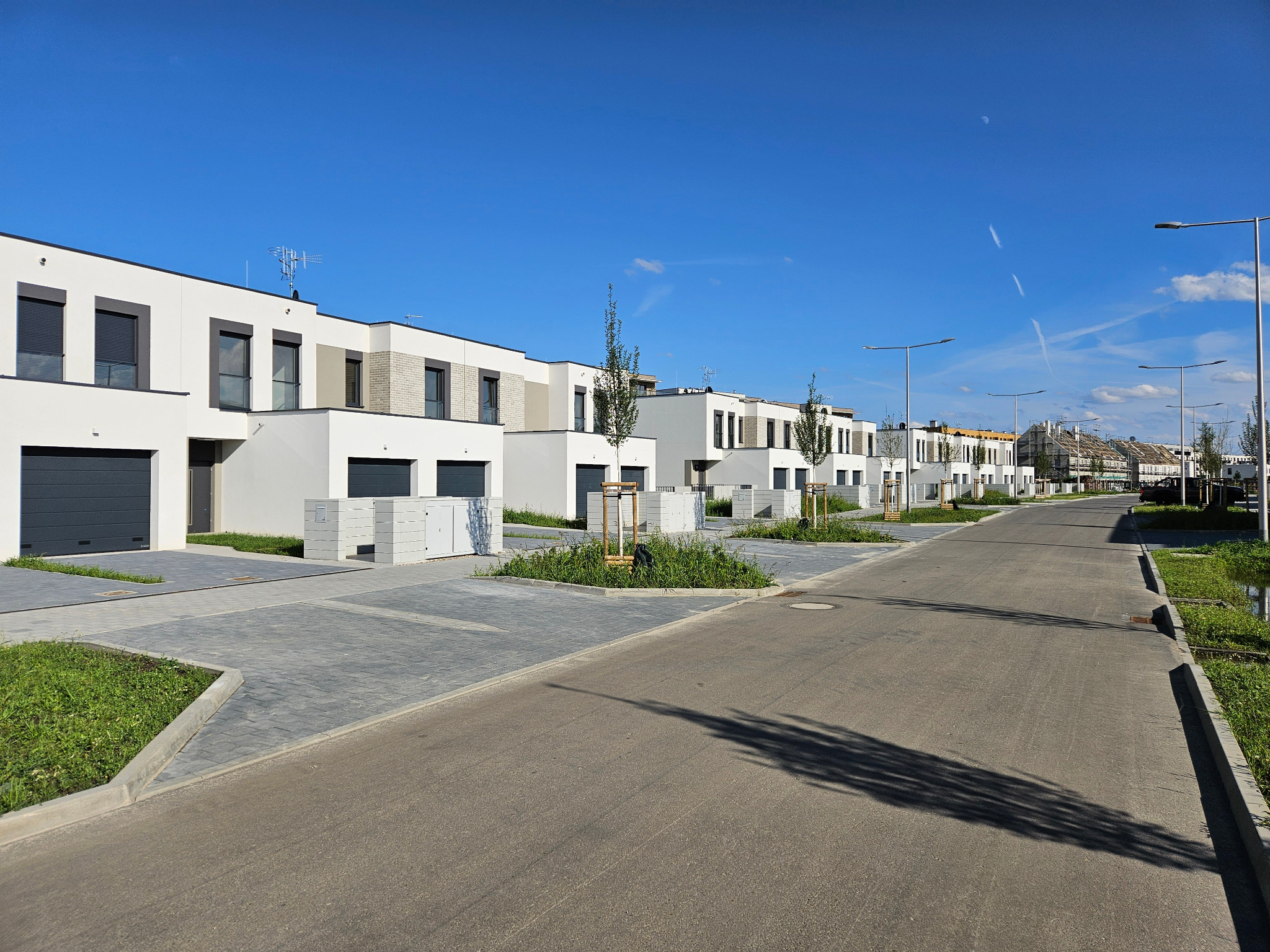 Nowoczesne osiedle domów szeregowych z płaskimi dachami, szarymi bramami garażowymi i kostką brukową na podjazdach, sfotografowane w słoneczny dzień.