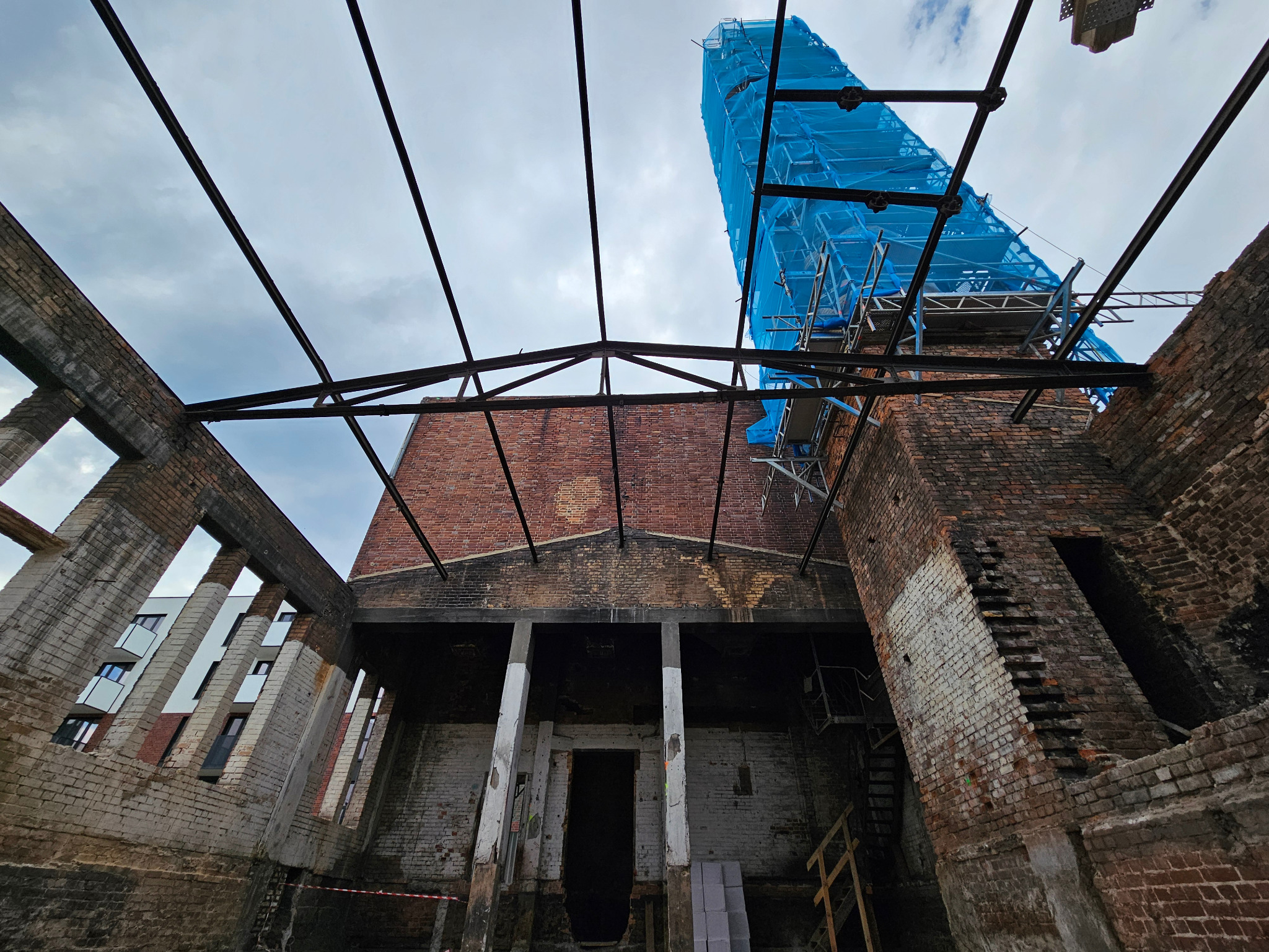 Wnętrze zrujnowanego budynku z cegły we Wrocławiu, z widoczną konstrukcją stalową dachu i rusztowaniami osłoniętymi niebieską siatką, ukazujące etap prac budowlanych.