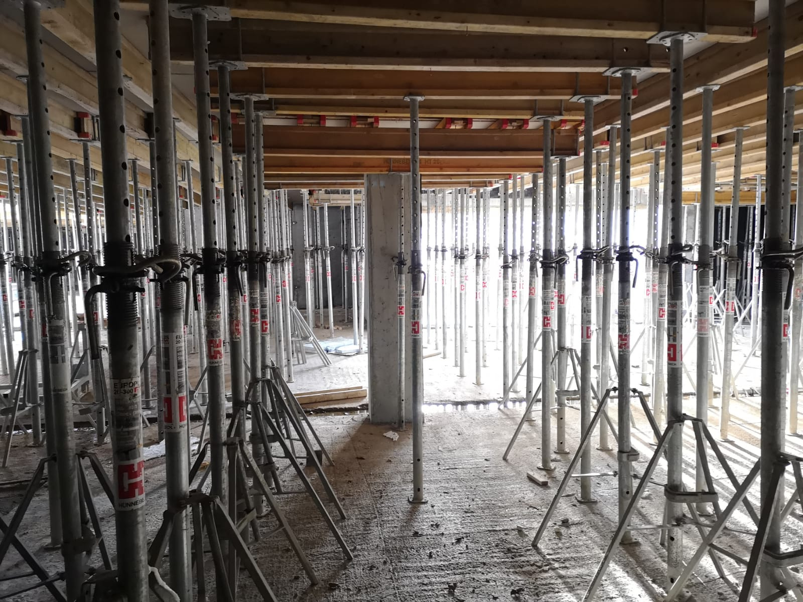 Szare stemple budowlane podpierające drewniany strop w surowym stanie, widoczne betonowe elementy konstrukcyjne.