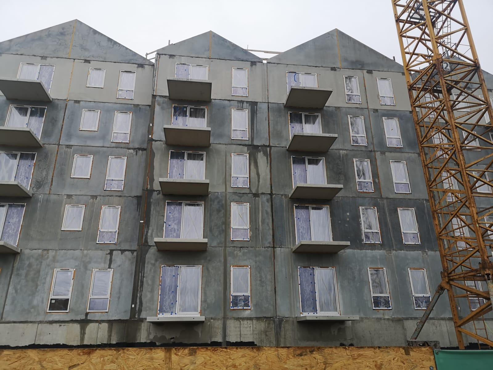 Surowy, betonowy budynek wielorodzinny w trakcie budowy, z widocznymi oknami zabezpieczonymi folią i fragmentem żółtego dźwigu budowlanego po prawej stronie.