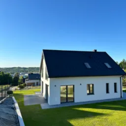 Nowoczesny dom jednorodzinny z płaskim, ciemnym dachem i białymi ścianami, otoczony świeżo założonym trawnikiem i widokiem na miasto w tle.