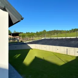 Betonowy fundament domu jednorodzinnego z izolacją przeciwwilgociową w postaci czarnej membrany, widok z poziomu trawnika w słoneczny dzień.