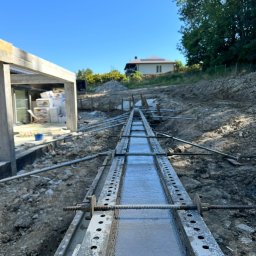 COMFORT INVEST SPÓŁKA Z OGRANICZONĄ ODPOWIEDZIALNOŚCIĄ - Wąski, zalany betonem szalunek muru oporowego, widoczny z perspektywy ziemi, z fragmentem budynku w tle i błękitnym niebem. Prace ziemne w toku.