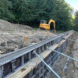 COMFORT INVEST SPÓŁKA Z OGRANICZONĄ ODPOWIEDZIALNOŚCIĄ - Wykop z szalunkiem wypełnionym betonem, w tle żółta koparka Liugong i drzewa. Widoczne metalowe podpory szalunku. Prace ziemne i betonowanie w toku.