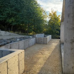 COMFORT INVEST SPÓŁKA Z OGRANICZONĄ ODPOWIEDZIALNOŚCIĄ - Betonowe mury oporowe w wykopie, wypełnione żwirem, z widocznym fragmentem konstrukcji budynku w tle i zielenią drzew. Ujęcie z poziomu gruntu, ciepłe światło.