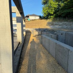 COMFORT INVEST SPÓŁKA Z OGRANICZONĄ ODPOWIEDZIALNOŚCIĄ - Betonowe mury oporowe w trakcie budowy, widoczny cień osoby, teren po niwelacji, w tle dom z panelami słonecznymi, błękitne niebo.