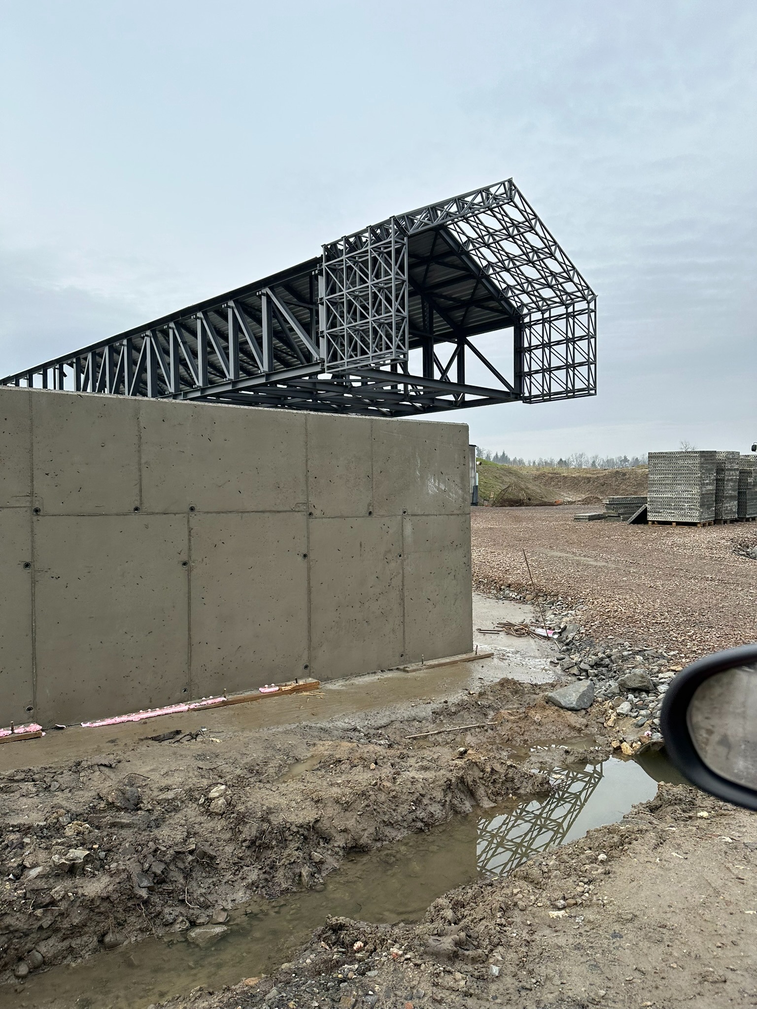Surowy betonowy fundament z widoczną izolacją termiczną i stalowa konstrukcja szkieletowa dachu w budowie, na tle gruntu z kałużami i paletą materiałów budowlanych.