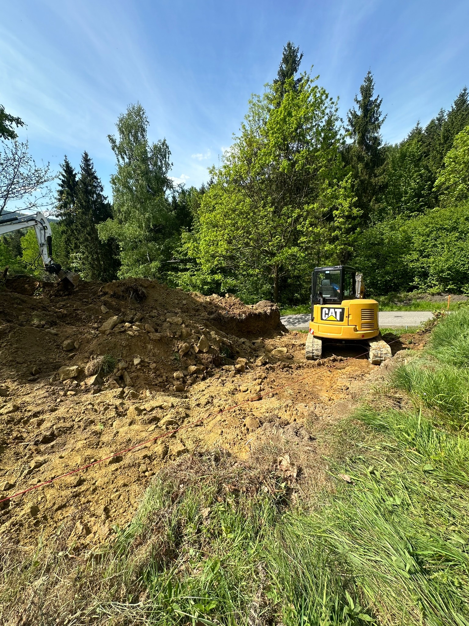 Żółta koparka CAT na terenie budowy, przygotowująca teren pod przyszłe prace, w tle zieleń drzew i częściowo widoczna druga koparka.