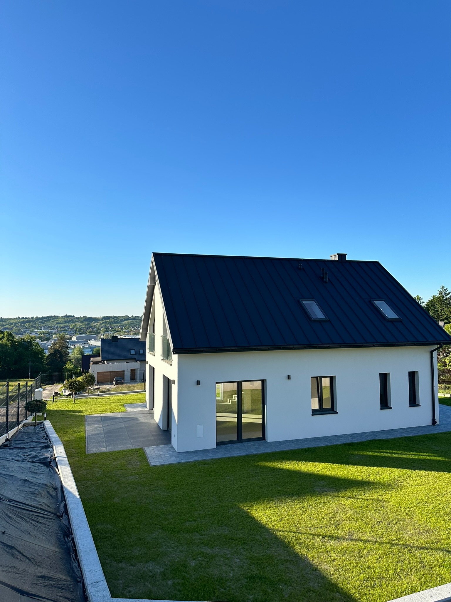 Nowoczesny dom jednorodzinny z płaskim, ciemnym dachem i białymi ścianami, otoczony świeżo założonym trawnikiem i widokiem na miasto w tle.