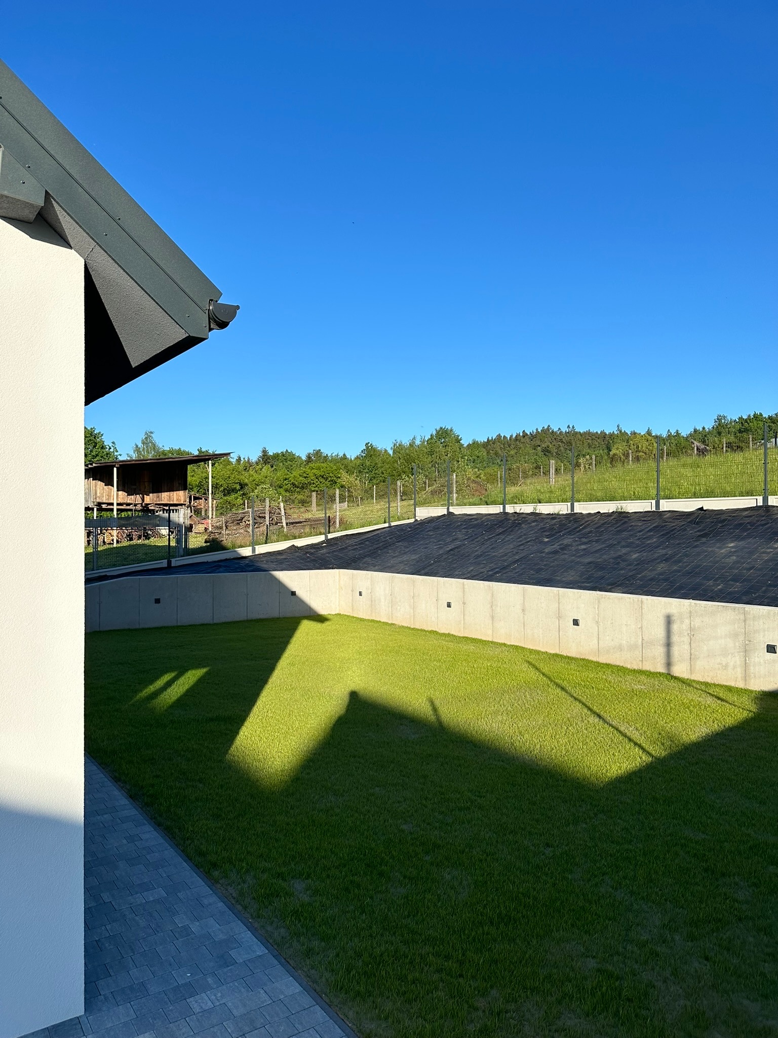 Betonowy fundament domu jednorodzinnego z izolacją przeciwwilgociową w postaci czarnej membrany, widok z poziomu trawnika w słoneczny dzień.
