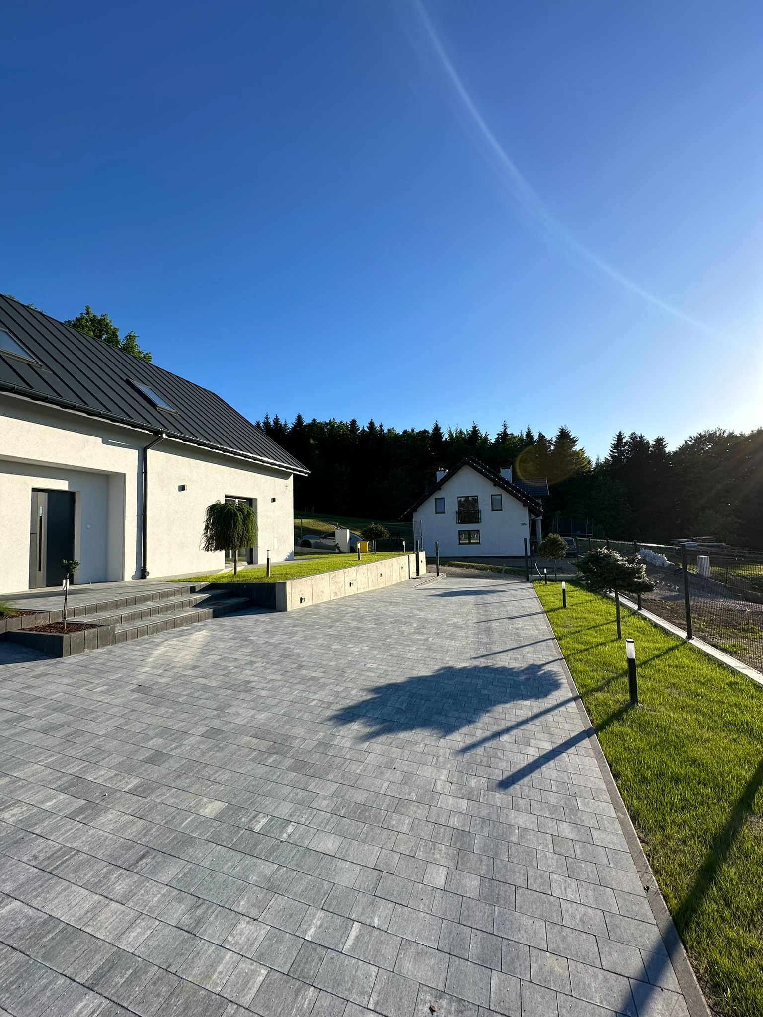 Nowoczesny dom jednorodzinny z jasną elewacją i ciemnym dachem, widok z brukowanego podjazdu w słoneczny dzień, z widocznymi elementami zagospodarowania terenu.