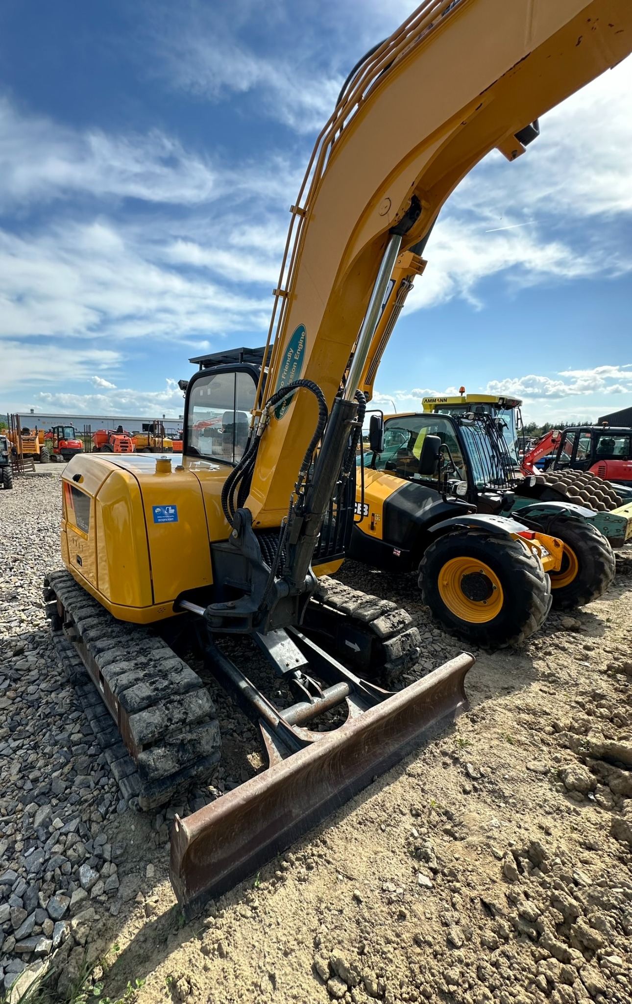 Żółta mini koparka gąsienicowa z lemieszem na żwirowym placu budowy, z innymi maszynami w tle i błękitnym niebem z chmurami.