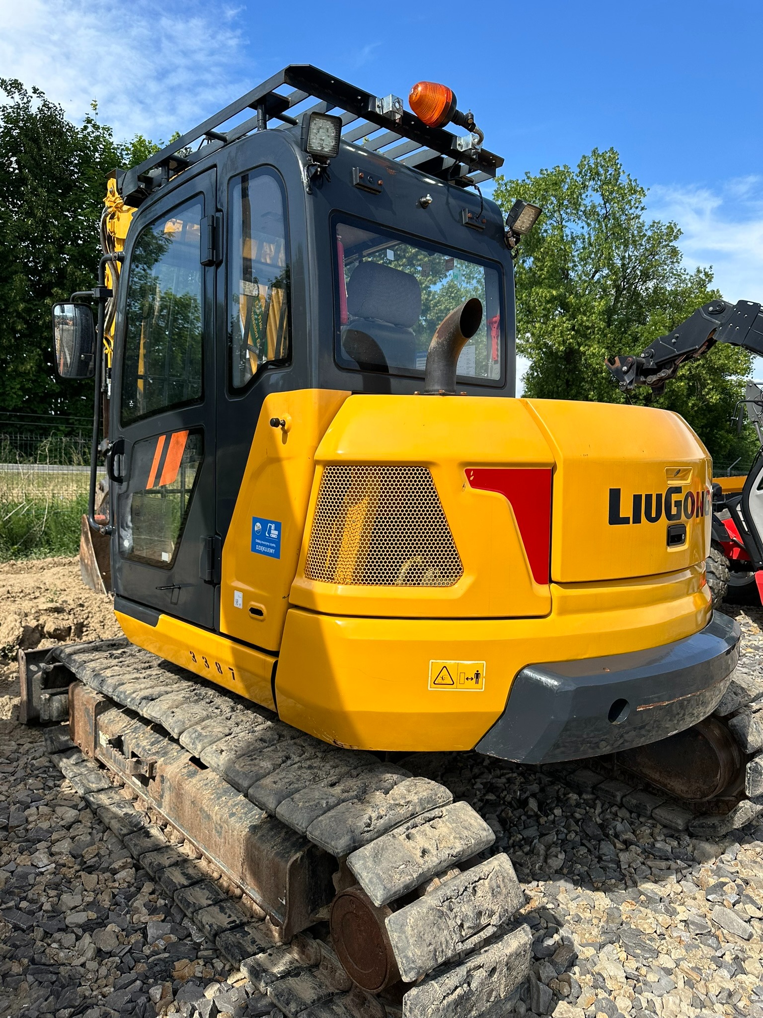 Żółto-szara mini koparka gąsienicowa marki LIUGONG, widoczna z tyłu, stojąca na żwirowym podłożu, z zielonymi drzewami w tle.