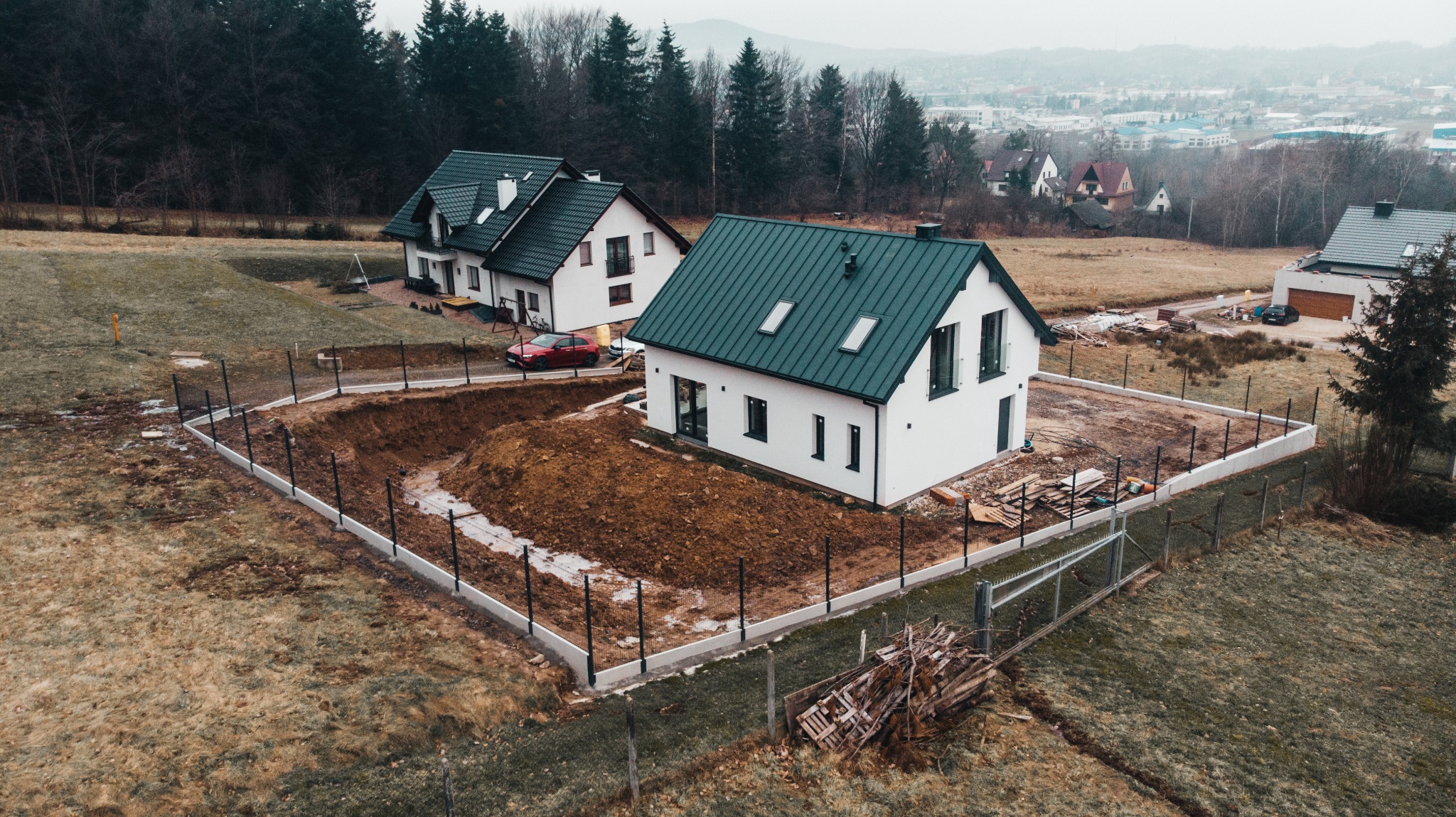 Widok z góry na dwa nowoczesne, białe domy z ciemnymi dachami; jeden z nich w trakcie budowy, otoczony wykopem pod fundamenty i ogrodzeniem, z widocznym stosem drewna budowlanego w rogu działki.